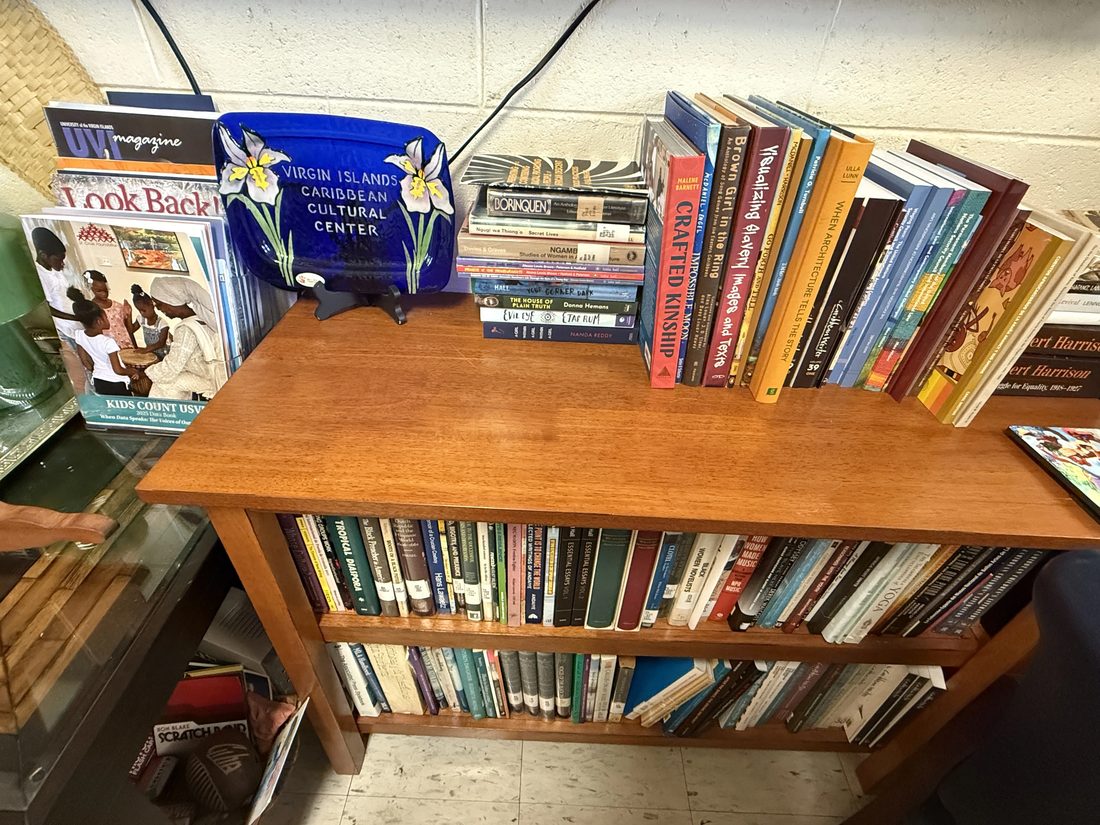 A wooden bookshelf topped with a VICCC serving tray and rows of Caribbean history books.